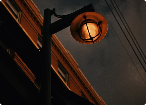 A streetlamp glows warmly at dusk, set against a darkening sky and the outline of a building, with power lines visible overhead.