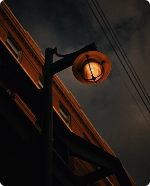 A streetlamp glows warmly at night, illuminating part of a building and power lines against a dark, cloudy sky.