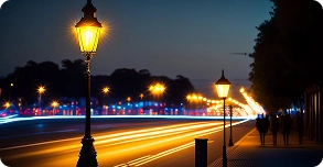 A city street at dusk with glowing streetlights, blurred car lights creating light trails, and a few people walking on the sidewalk lined with trees.