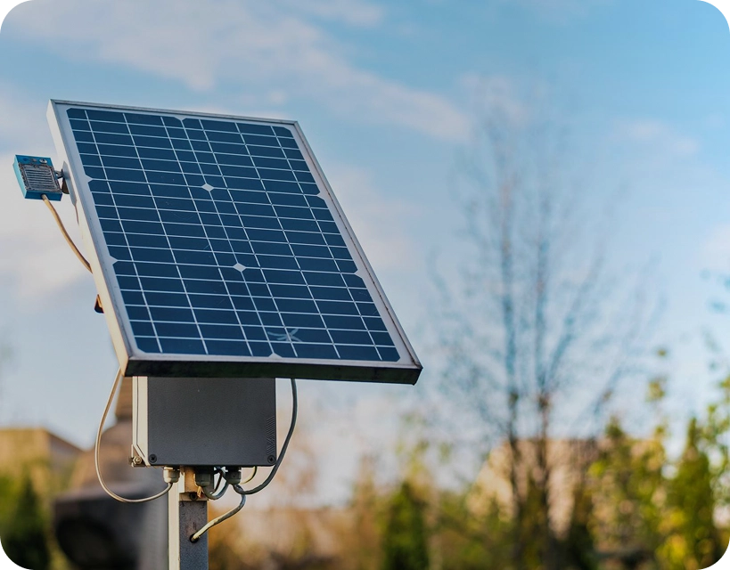 A solar panel mounted on a metal pole outdoors, with clear blue sky and blurred greenery in the background.