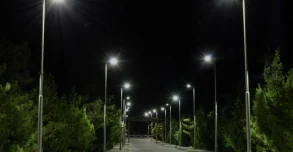 A paved road at night lined with tall streetlights on both sides, illuminating the path, with dense green trees visible along the edges.